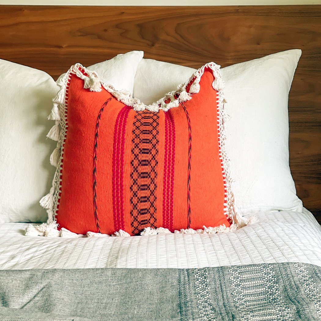 Colorful Throw Pillow With Fringe | Multiple Colors - BAJA CHICNEWorangeHUNTEDFOX - Bright boho pillow with fringe in orange, purple, and red tones on a modern bed styled with cozy decor, including a textured blanket.