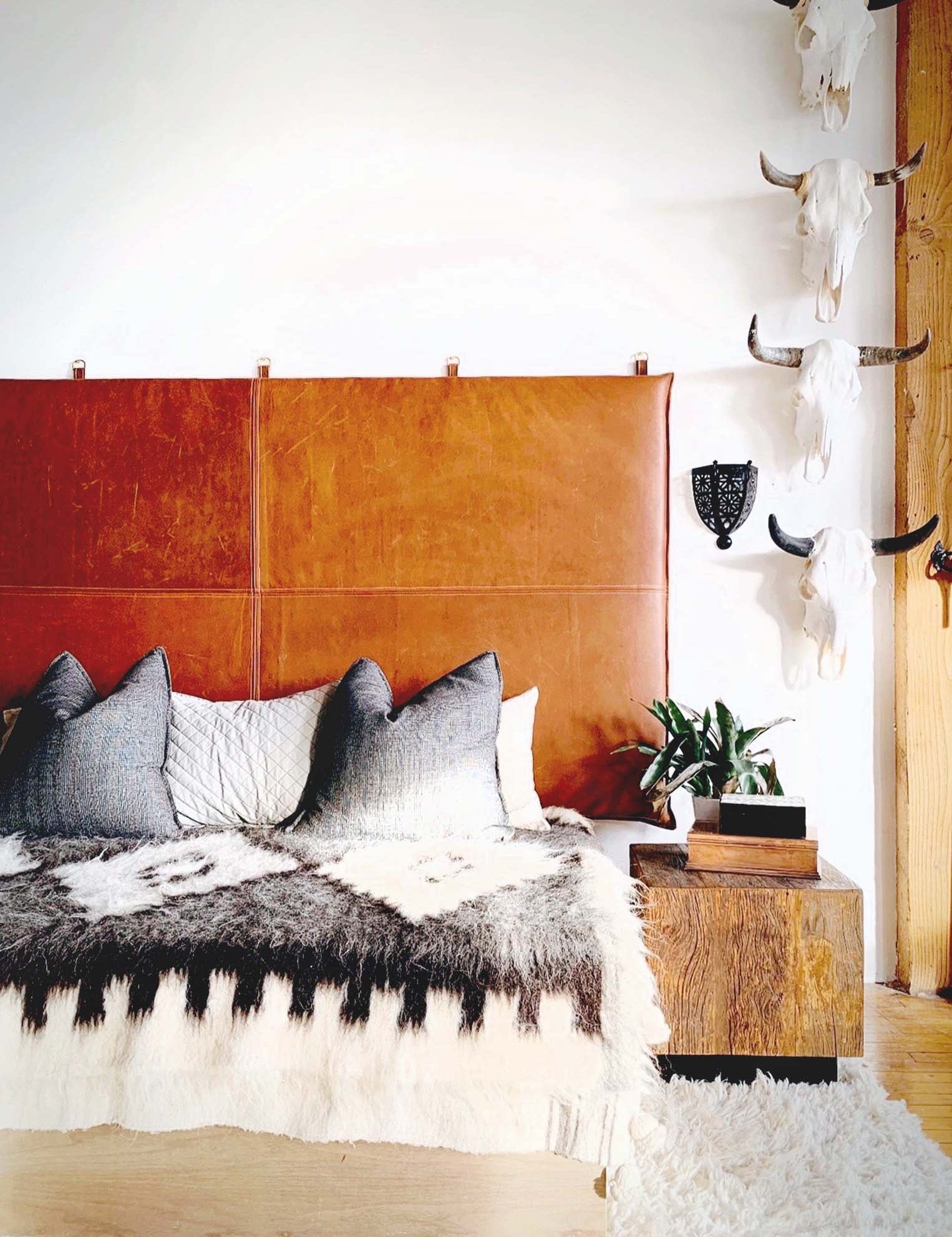 A large king-sized brown leather hanging headboard with white top stitching, mounted on a wall with decorative pillows on the bed. The room has a rustic and modern aesthetic with wooden elements and a white carpet.