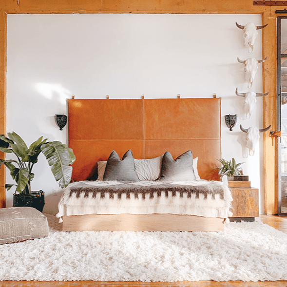 A large king-sized brown leather hanging headboard with white top stitching, mounted on a wall with decorative pillows on the bed. The room has a rustic and modern aesthetic with wooden elements and a white carpet.