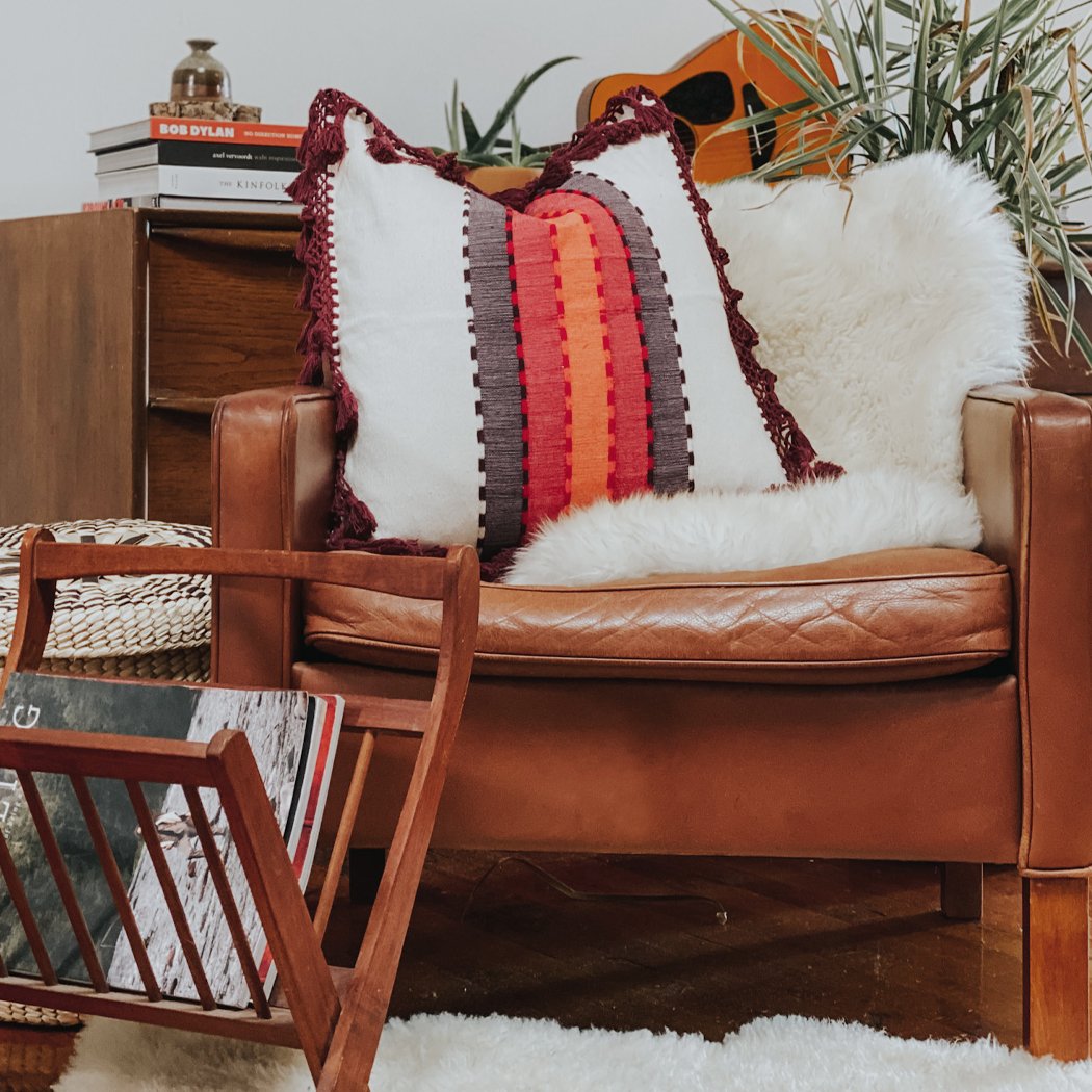 Bright boho pillow with fringe in orange, purple, and red tones on a leather chair, styled with cozy decor, including a sheepskin throw, plants, and wooden accents