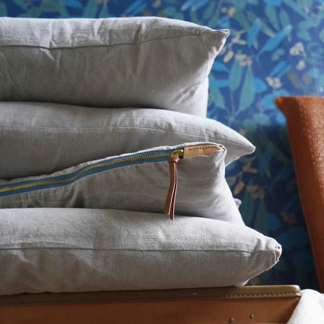 A stack of light grey accent pillows with a visible turquoise and brass zipper on one of the pillows, placed on a wooden surface with a patterned wallpaper in the background.