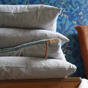 A stack of light grey accent pillows with a visible turquoise and brass zipper on one of the pillows, placed on a wooden surface with a patterned wallpaper in the background.