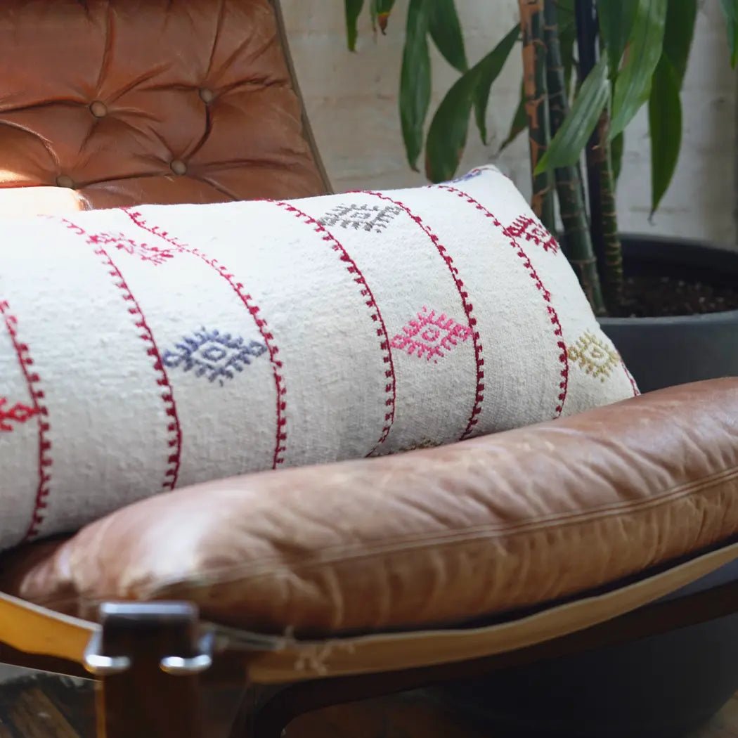 Handwoven cotton lumbar pillow with red and blue embroidery on an ivory base, styled on a leather armchair.