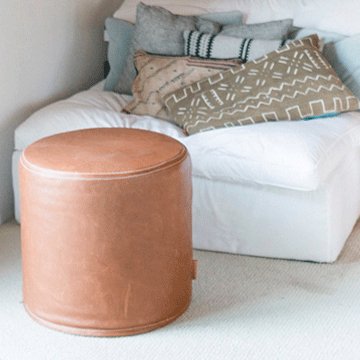 brown round leather ottoman sitting in front of a white sofa with geometric pillows sitting on top