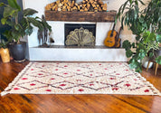 Handwoven Moroccan-style area rug with abstract village motifs in vibrant colors, including pink, turquoise, and black, on a cream background with fringe detailing. Styled in a cozy in a fireplace setting.   