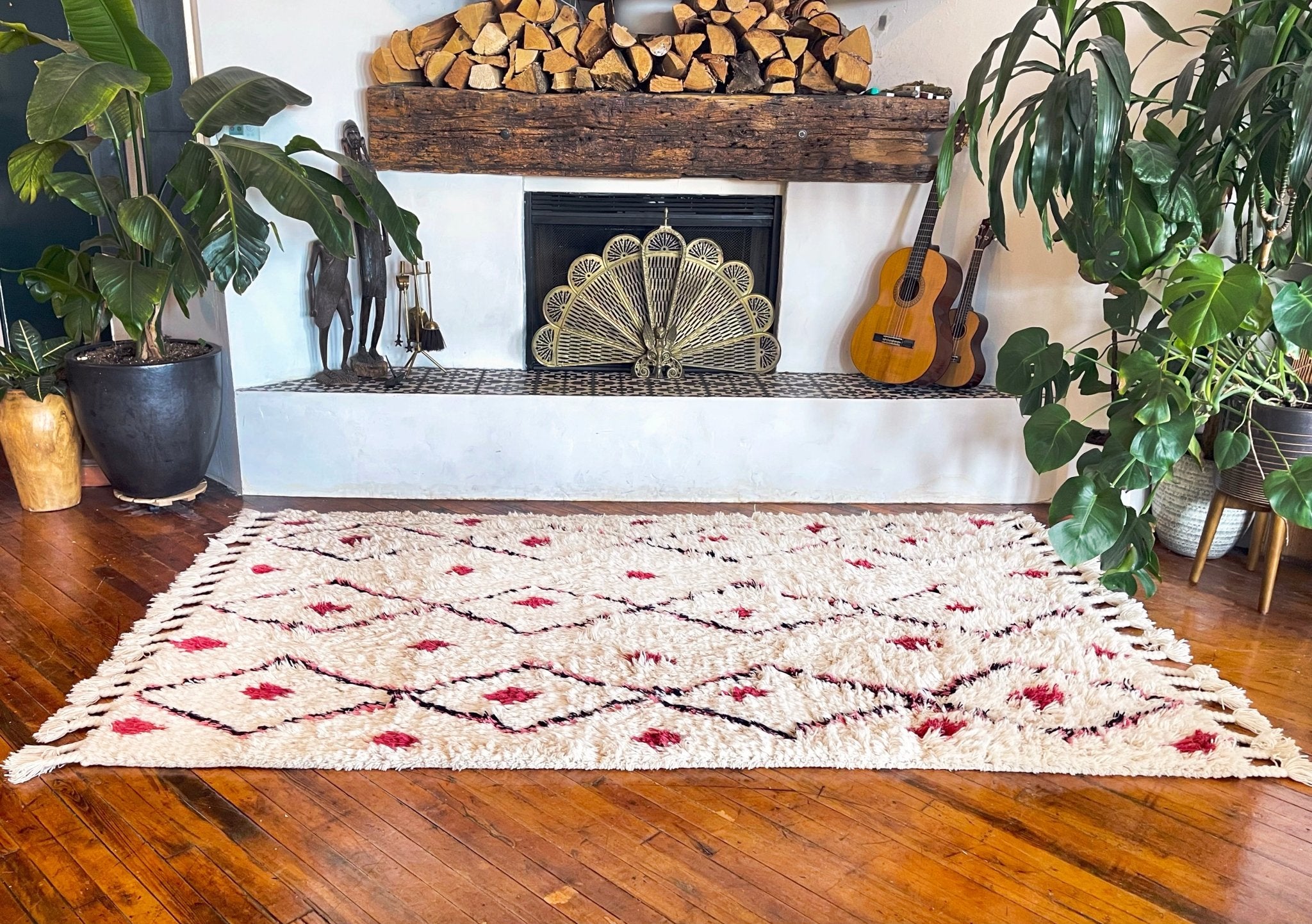 Handwoven Moroccan-style area rug with abstract village motifs in vibrant colors, including pink, turquoise, and black, on a cream background with fringe detailing. Styled in a cozy in a fireplace setting.   